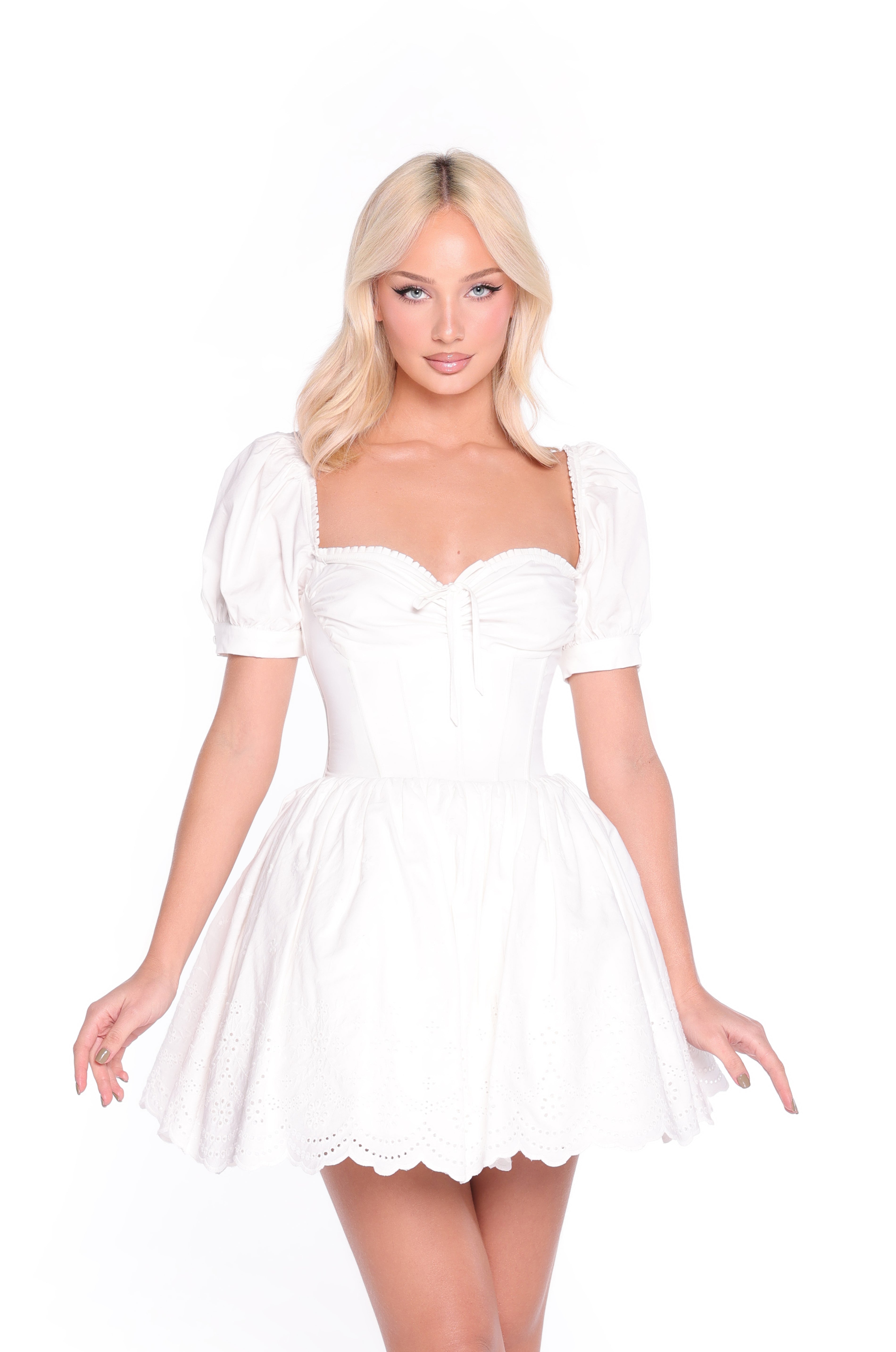 Woman wearing the Celine Fit-and-Flare Short White Grad Dress, featuring puff sleeves, a fitted bodice, and an eyelet flared skirt, standing against a plain white background.